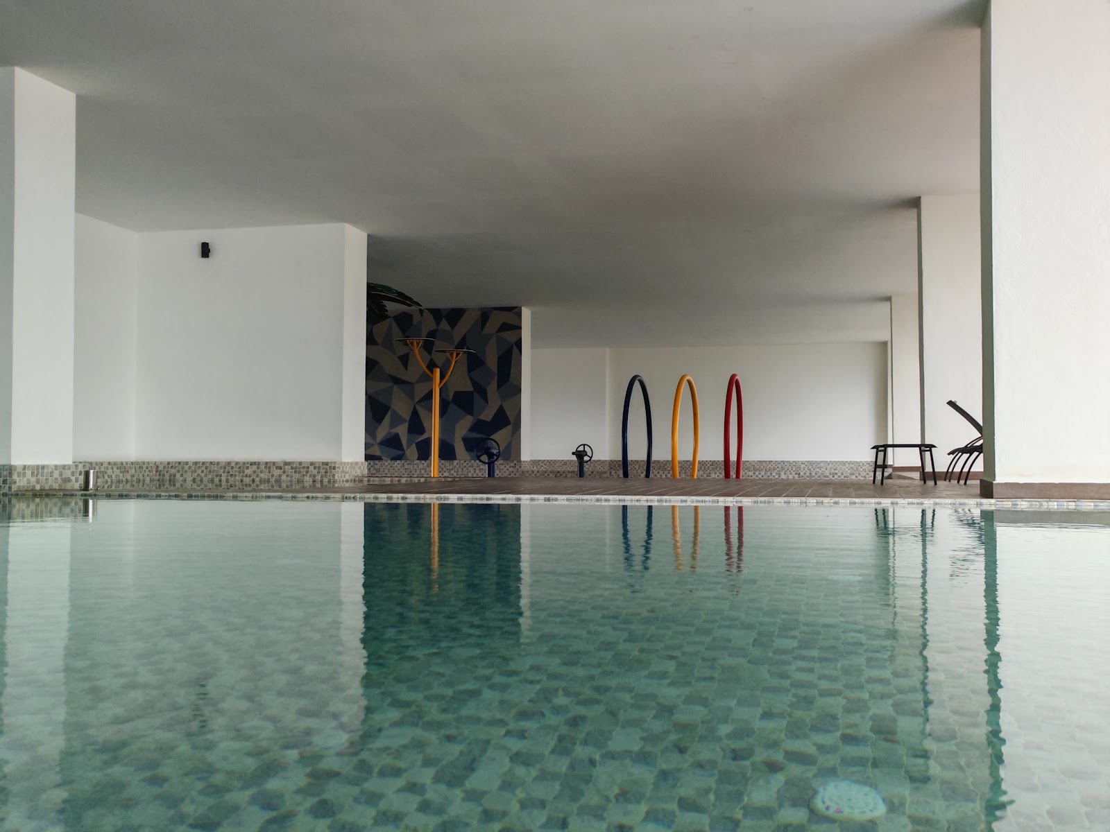 Residensi Bintang Bukit Jalil: A calm swimming pool with three colorful arched sculptures standing on the edge, surrounded by white walls.