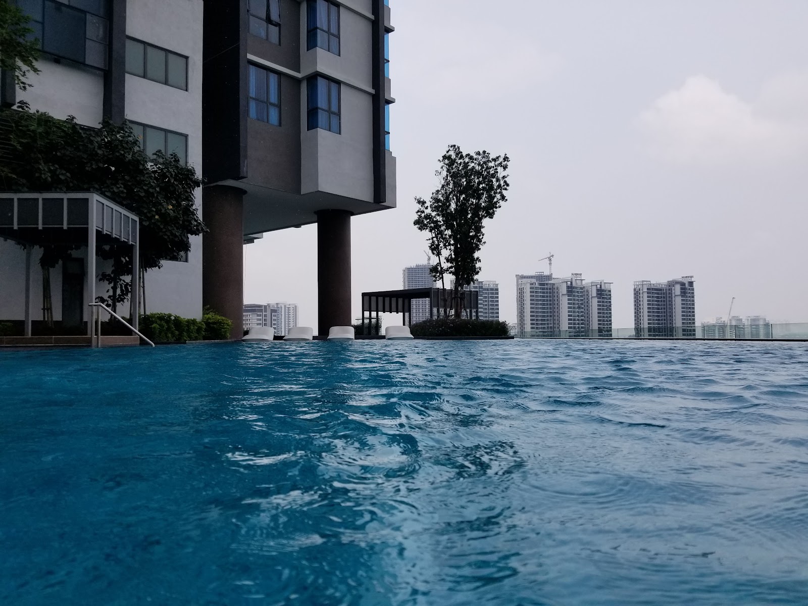 SUNWAY GEOSENSE: An elevated swimming pool with blue water and white loungers on a rooftop, overlooking a cityscape with high-rise buildings under construction in the background on an overcast day.