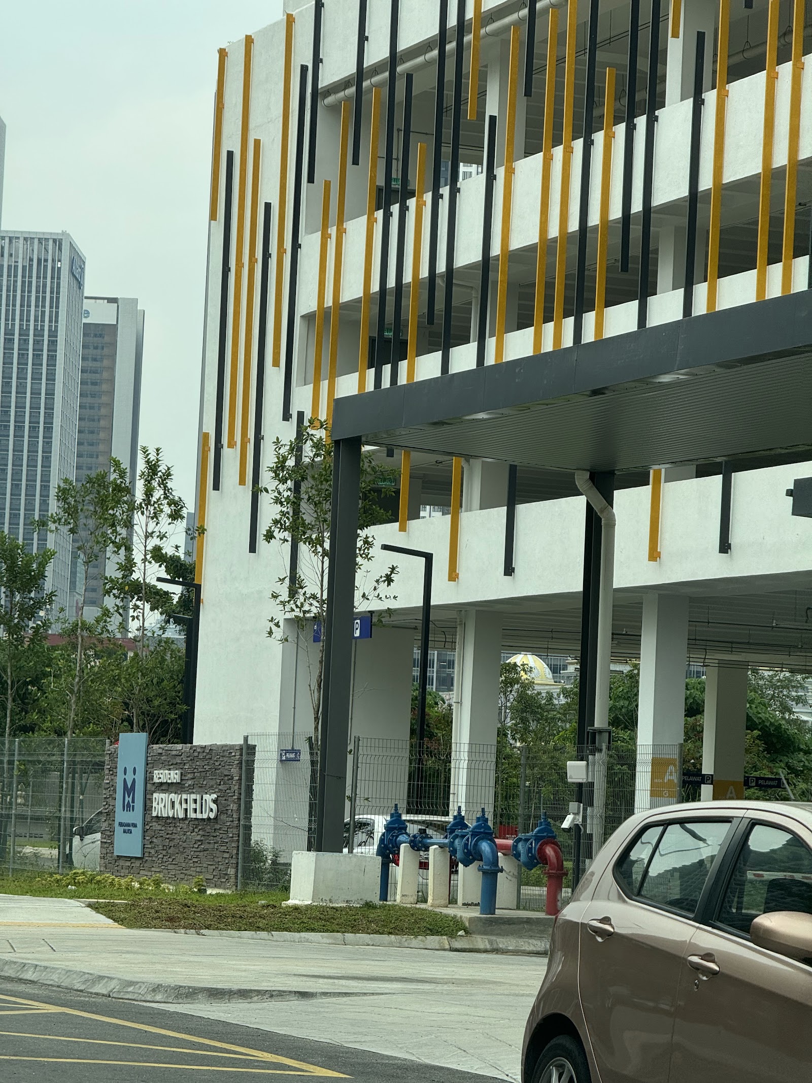 RESIDENSI BRICKFIELDS: A photograph of the exterior of the Brickfields residence, showing its modern facade with vertical yellow and black accents, the entrance area, and a project sign.