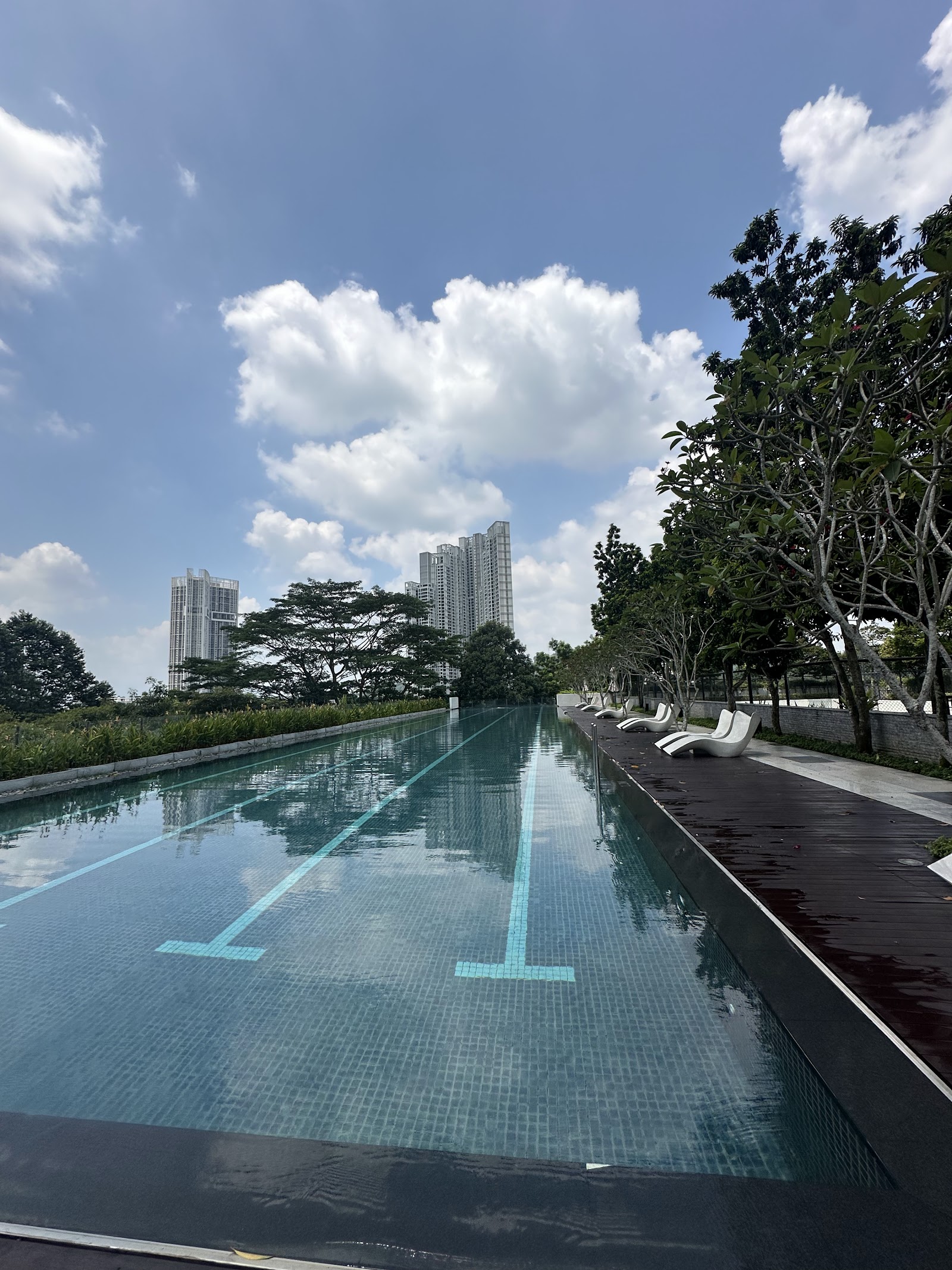 One Central Park @ Desa ParkCity: A long swimming pool on a rooftop or terrace, featuring lane markers, white sun loungers, and a backdrop of trees and high-rise residential buildings under a cloudy sky.