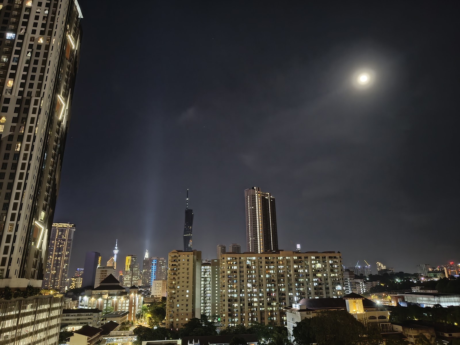 The Pines: A night view from a high-rise balcony overlooking a city skyline with a bright full moon in the sky.