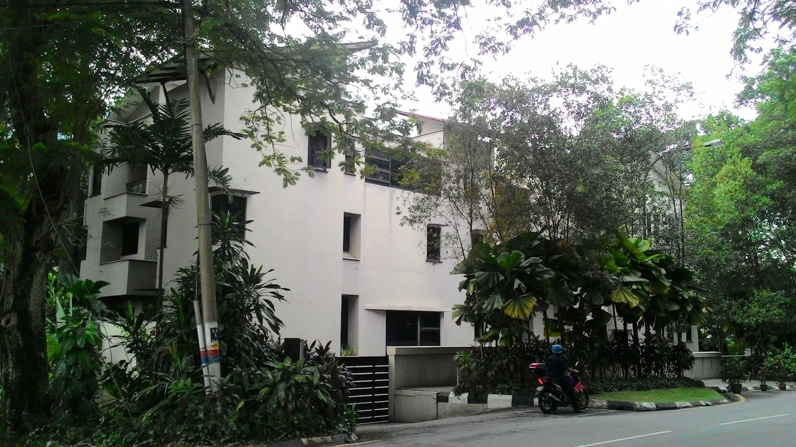 78 LAD: A white multi-story residential building surrounded by trees and landscaping with a motorcyclist on the adjacent road.