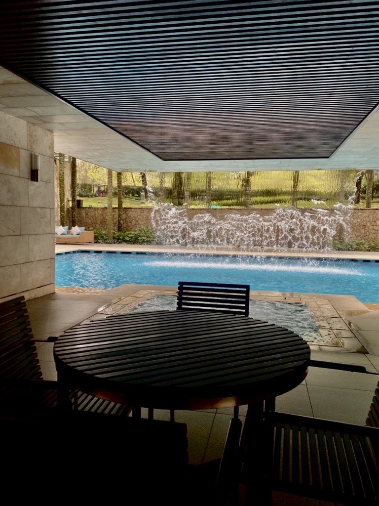 The Binjai On The Park: A covered seating area with a round table and chairs, overlooking a swimming pool with a cascading waterfall feature and garden in the background.