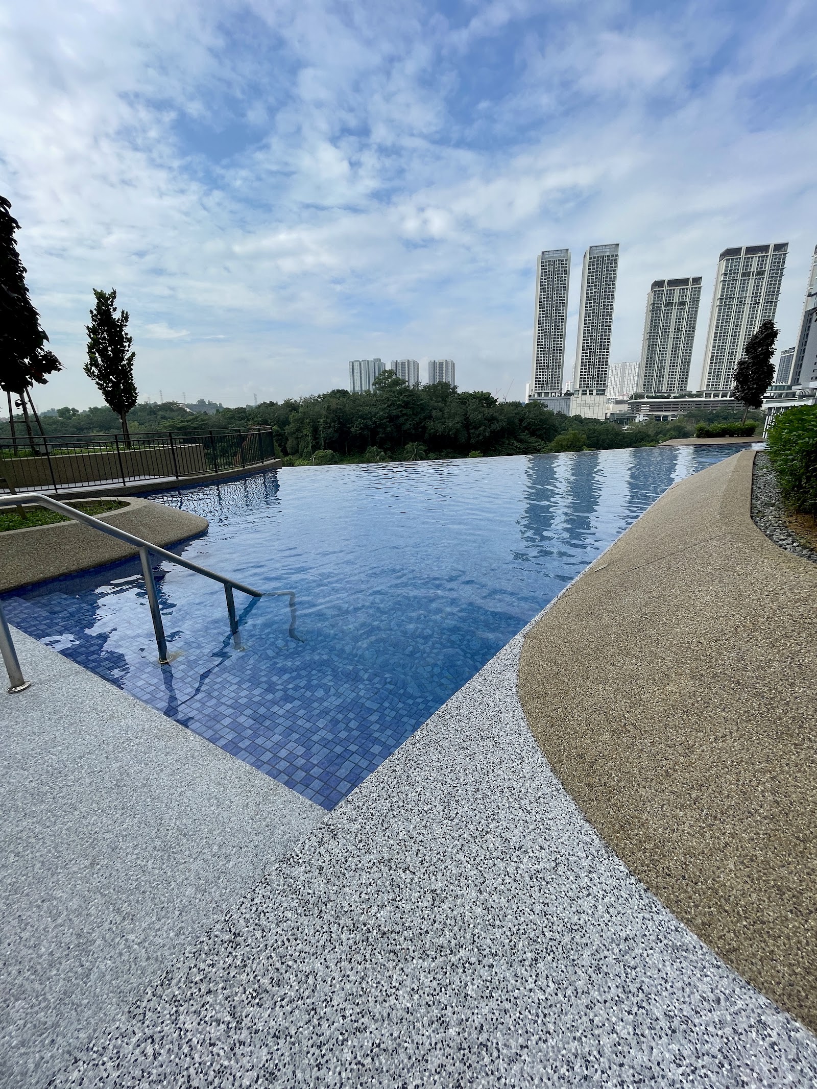 The Tropika Bukit Jalil: An outdoor infinity swimming pool with a metal ladder, surrounded by a speckled pool deck and set against a backdrop of greenery and high-rise condominiums.