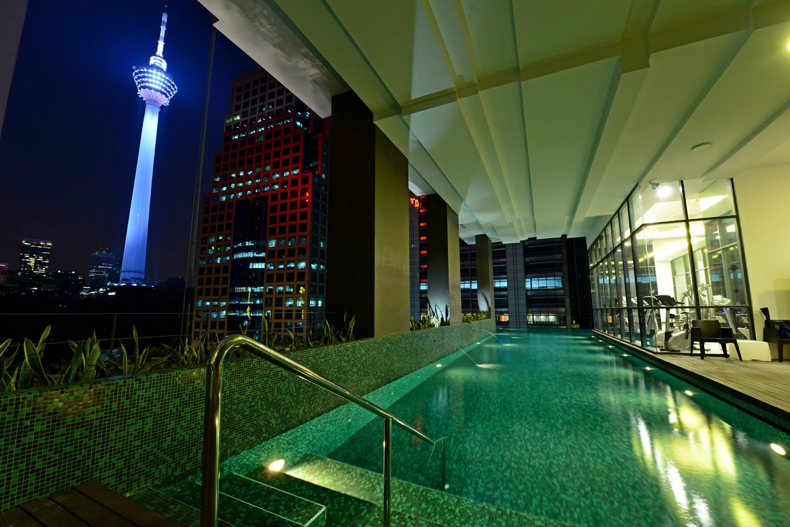 The Manhattan: A lit, tiled swimming pool with a glass-walled gym to the right, overlooking a night cityscape with a prominent tower.