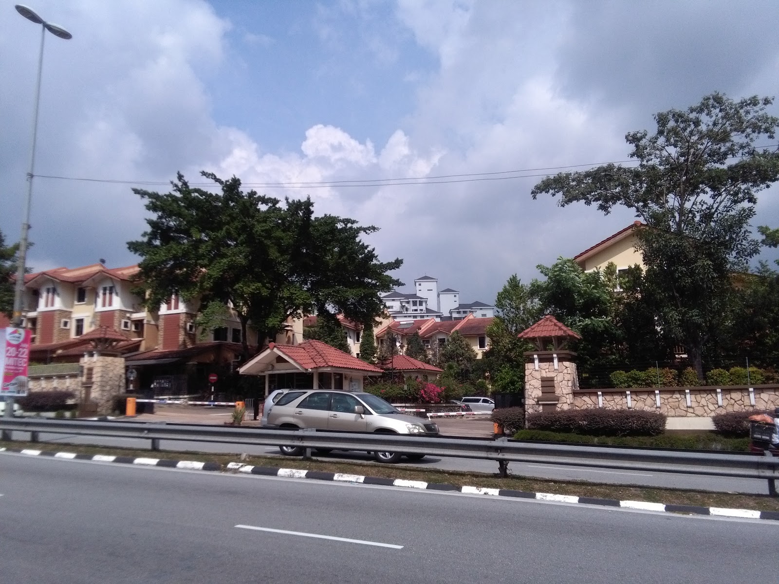 Prima Duta: A street-level view looking into a residential development, showing the entrance gate with stone pillars and a guard house, with townhouses and cars in the background.