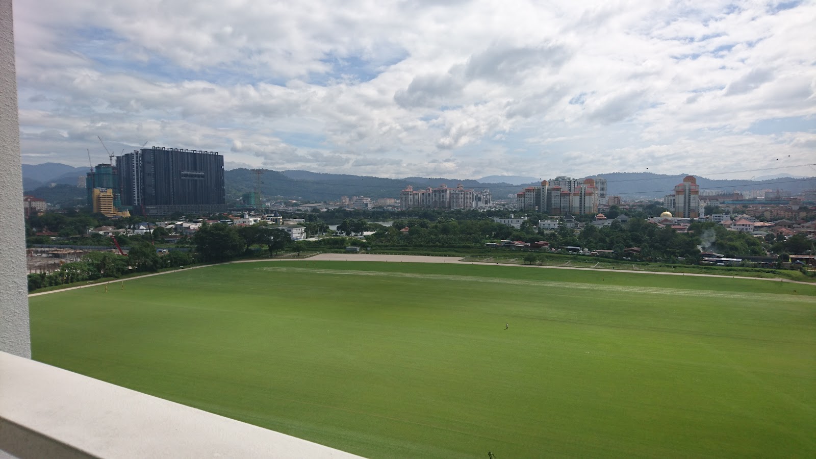 Mutiara Upper East: A view from a balcony overlooking a large green sports field with a distant city skyline and mountains in the background.