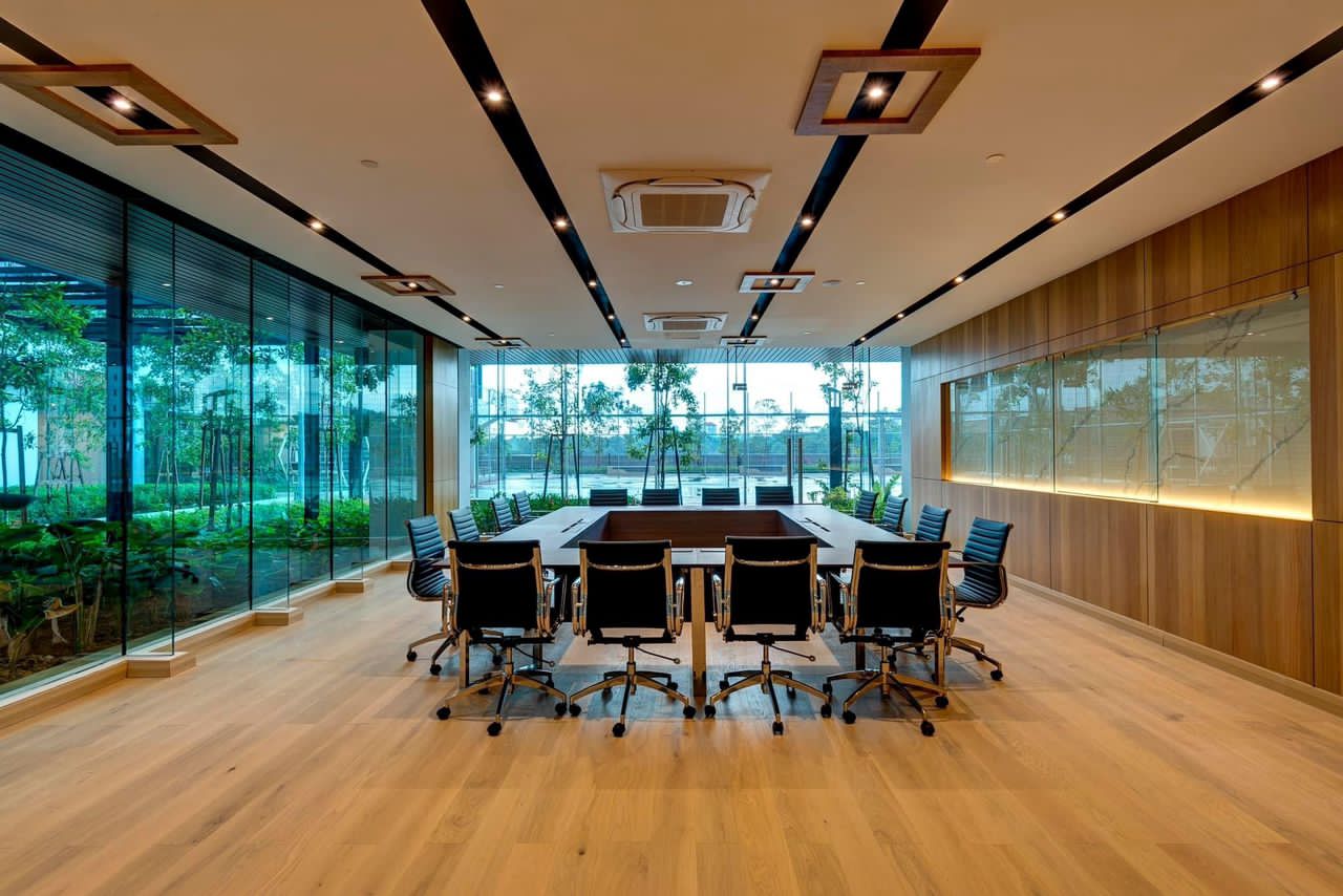 Sentral Suites: A spacious conference room with a long table, black office chairs, wooden flooring, and large glass windows overlooking a landscaped garden.