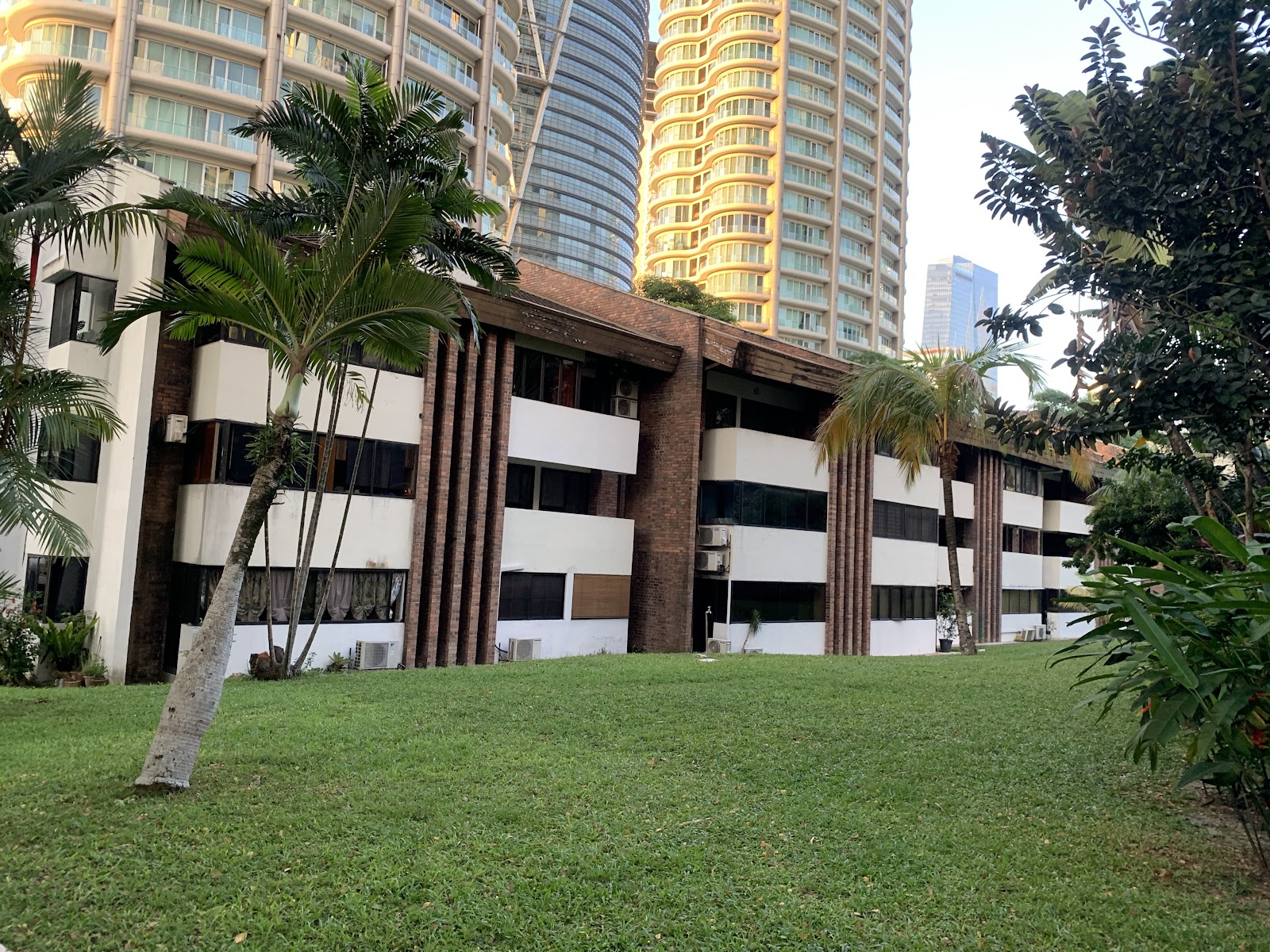 Desa Kudalari: The exterior of a multi-story building with a brick and white facade, viewed from across a manicured lawn with palm trees, with tall high-rise buildings in the background.