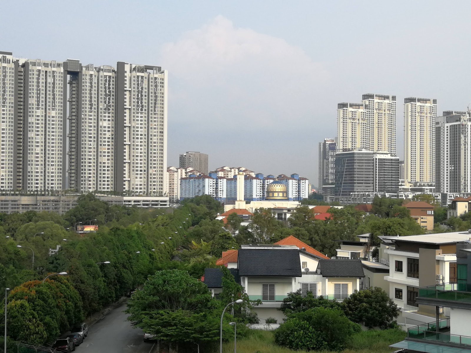 Denai Sutera: A distant view from a property overlooking a mix of low-rise terraced houses in the foreground, a mosque with a golden dome in the mid-ground, and a dense skyline of modern high-rise condominium towers in the background, set against a hazy sky.
