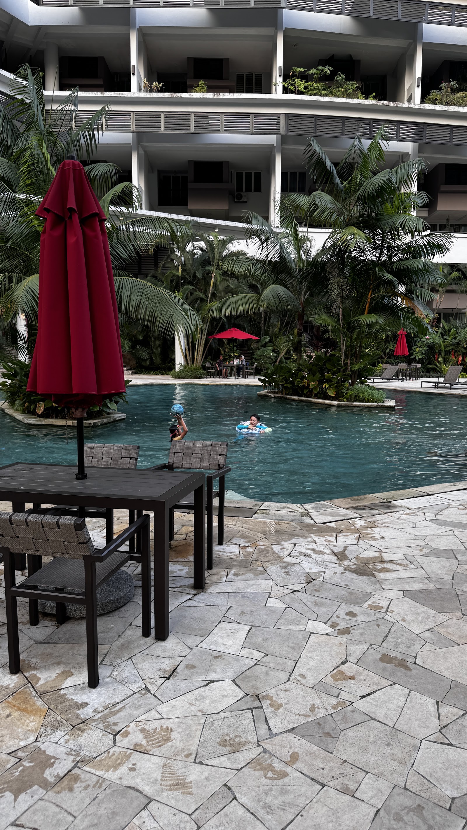 Armanee Terrace: A swimming pool area at a resort with people swimming, surrounded by lush tropical plants and a multi-story building in the background.