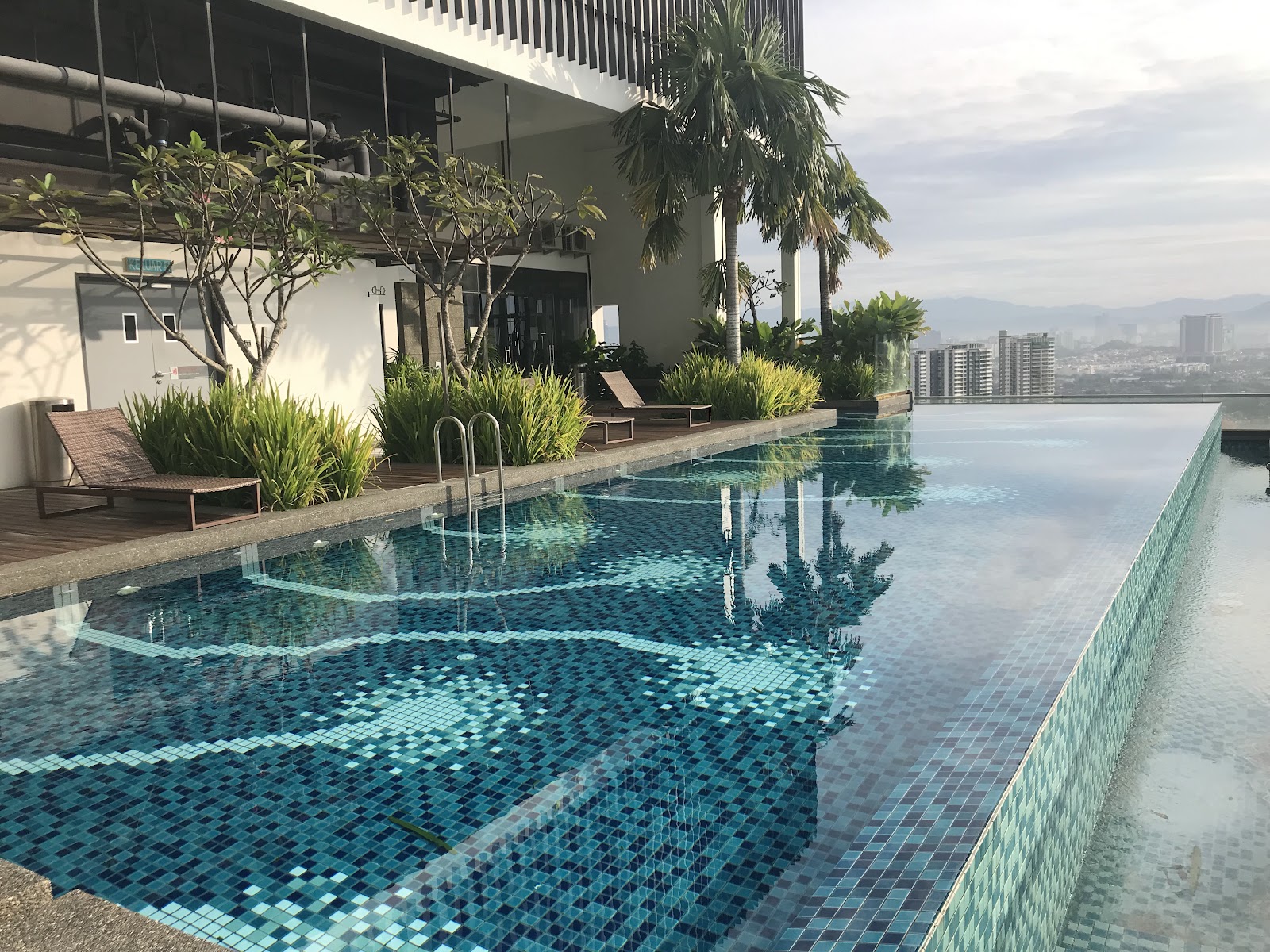 Rev0 @ Aurora Place: A rooftop infinity pool with mosaic tiles, sun loungers, and tropical landscaping on a wooden deck, overlooking a city and mountains in the distance.