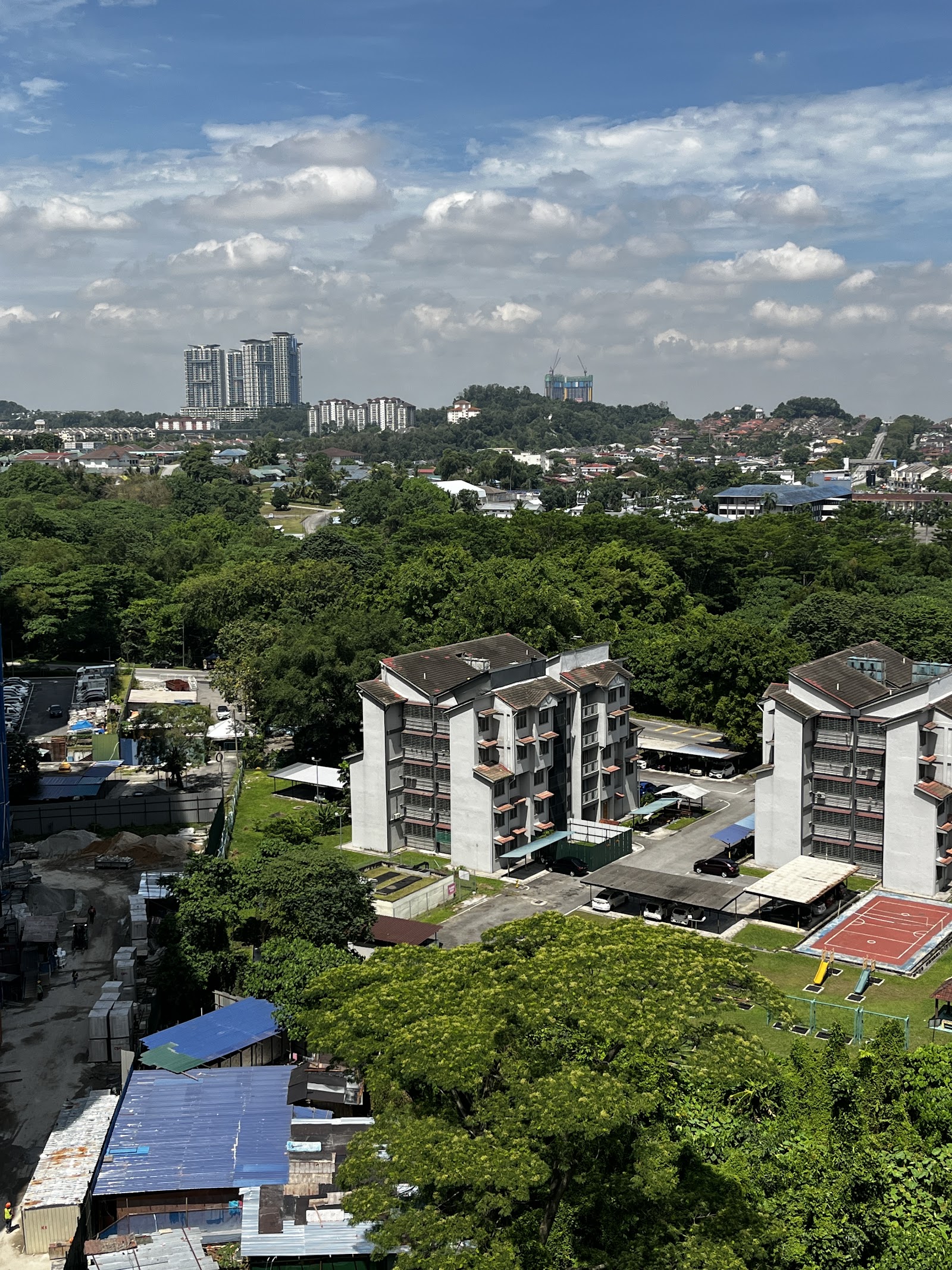 Kinrara Mas: An elevated view from a high-rise looking out over a residential area with apartment blocks and dense greenery, with a city skyline in the distance under a partly cloudy sky.