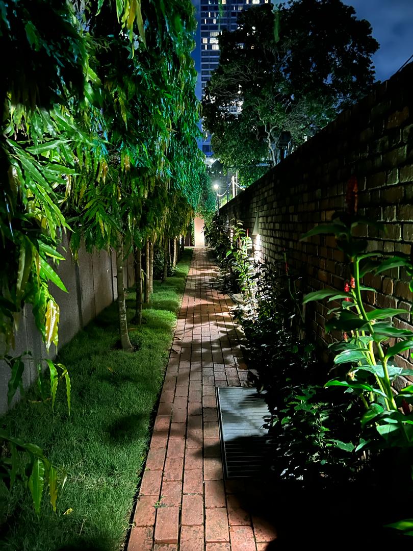 Dedaun Condominium: A paved pathway lined with trees, grass, and low shrubbery runs through a landscaped garden area at night, enclosed by walls.