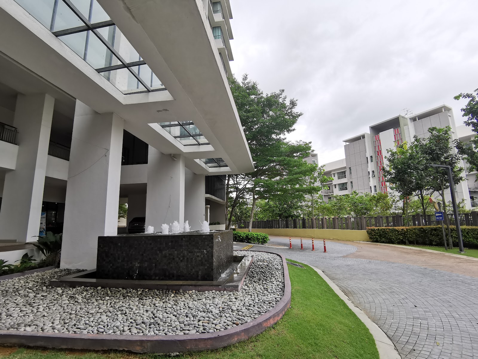 KM1: A landscaped courtyard area within a modern residential development, featuring a black stone fountain, paved walkways, and multi-story white apartment buildings under an overcast sky.