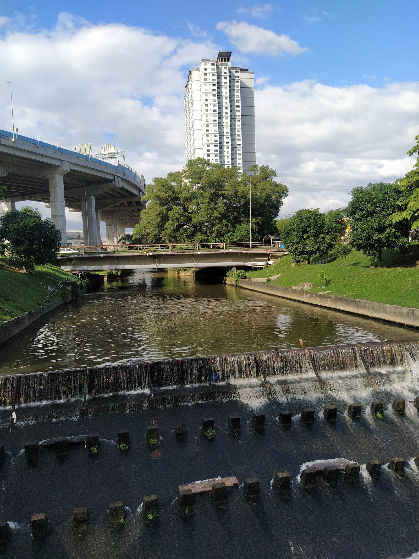 Casa Residenza @ Kota Damansara: A view down a canal with a small dam in the foreground, looking past a bridge towards a large white residential tower and a curved elevated highway.