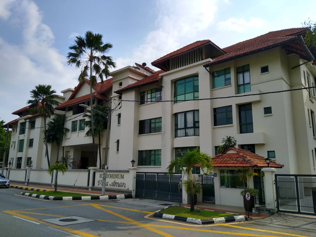 Kondominium Villa Aman: A street-level view of the white facade of Kondominium Villa Aman, showing its multi-story balconies, red tiled roof, and entrance gate with a security booth, set against a partly cloudy sky with palm trees in the foreground.