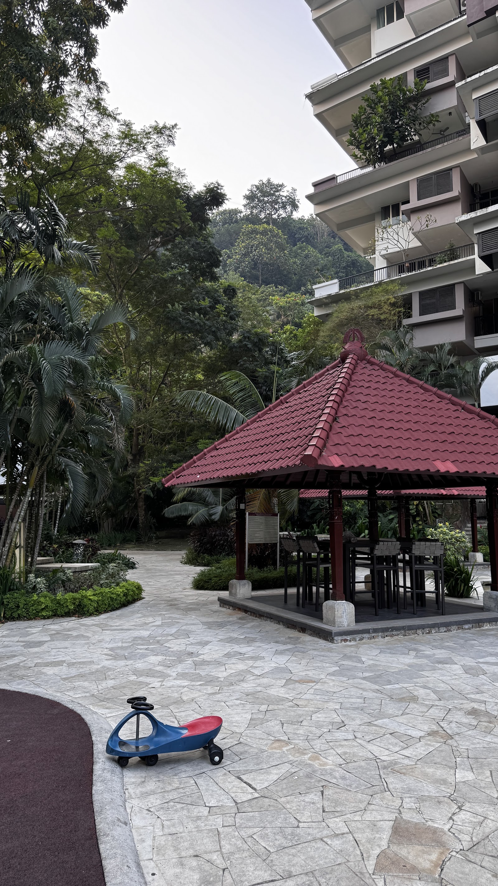 Armanee Terrace: A red-roofed gazebo with outdoor seating and a children's scooter on a stone-paved walkway in a landscaped garden area of a residential development.