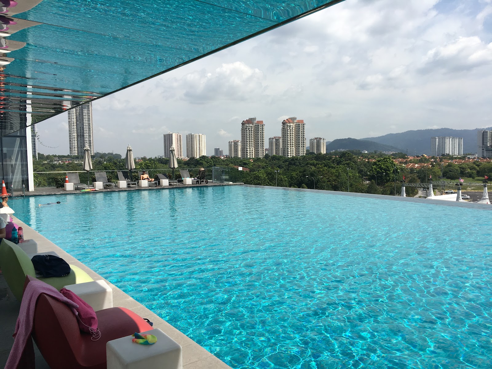 The BreezeWay Garden: A high-angle view of a rooftop infinity pool with lounge chairs, overlooking a cityscape with skyscrapers and a mountain range.
