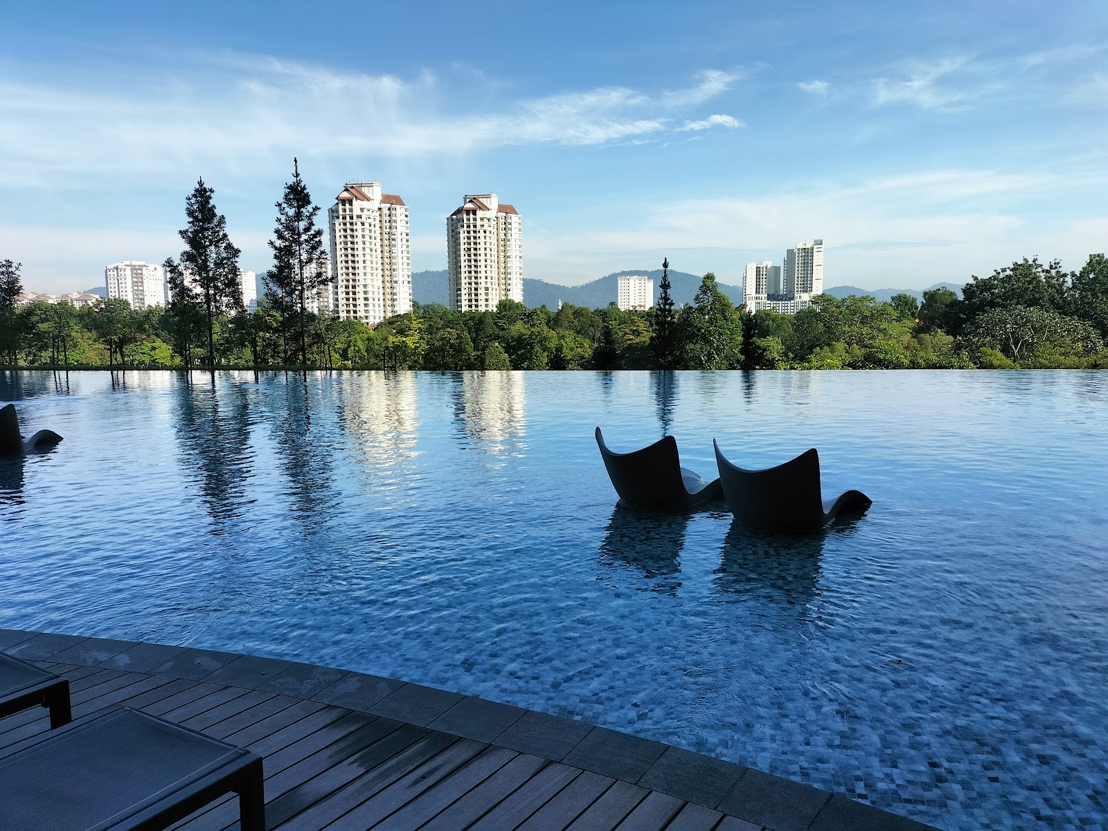 Park Regent by the Water: An infinity pool with modern water loungers, viewed from a wooden deck with high-rise buildings and trees in the background.