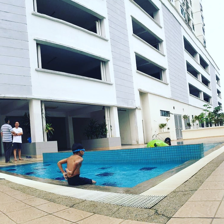 Kasturi Idaman: A man in swimwear sits in a blue tiled swimming pool with a green toy float, while two men stand on the deck near the building facade in the background.
