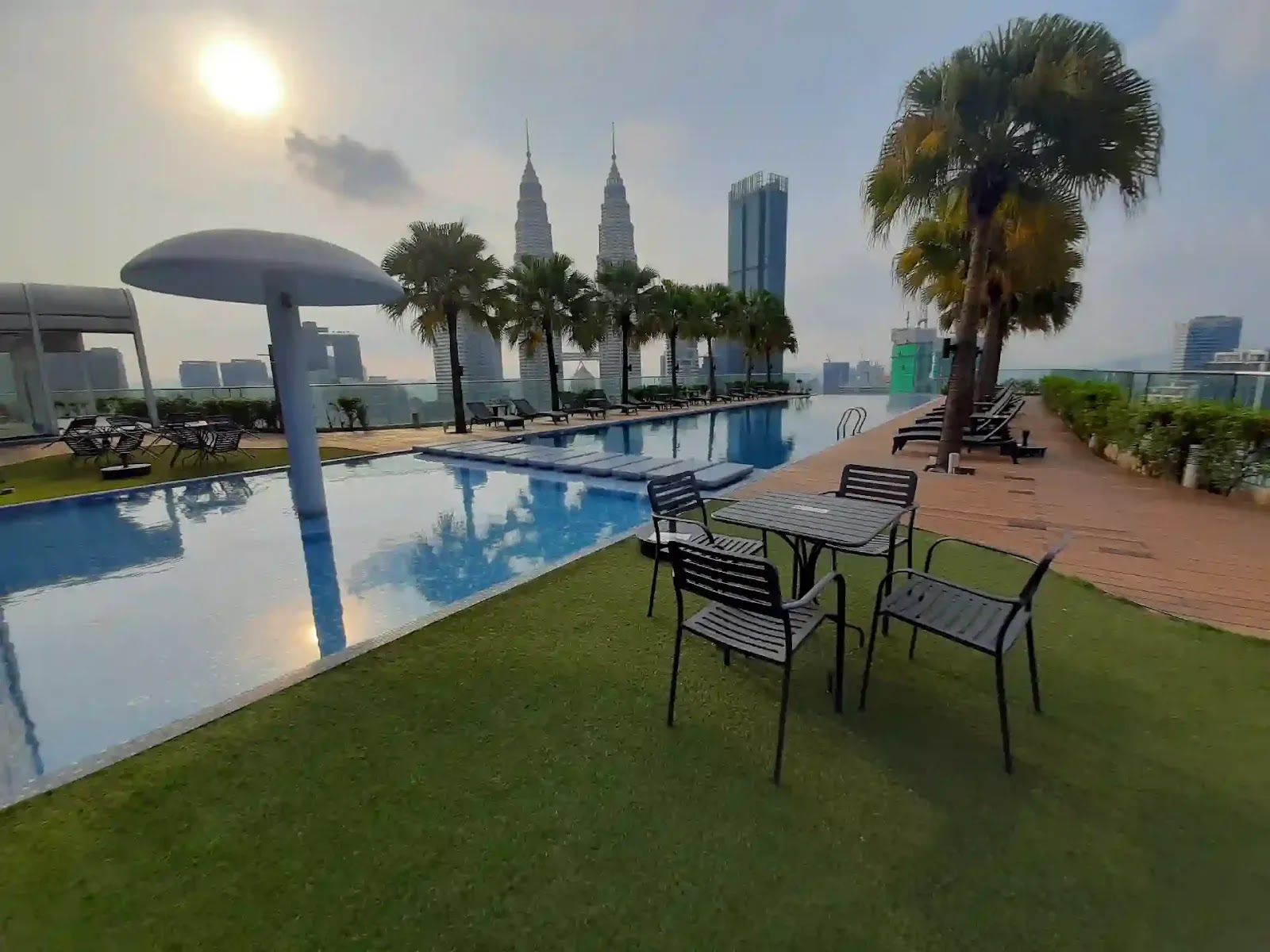 Quadro Residences: A rooftop swimming pool area with lounge chairs and seating on artificial grass, overlooking the Kuala Lumpur city skyline including the Petronas Twin Towers.