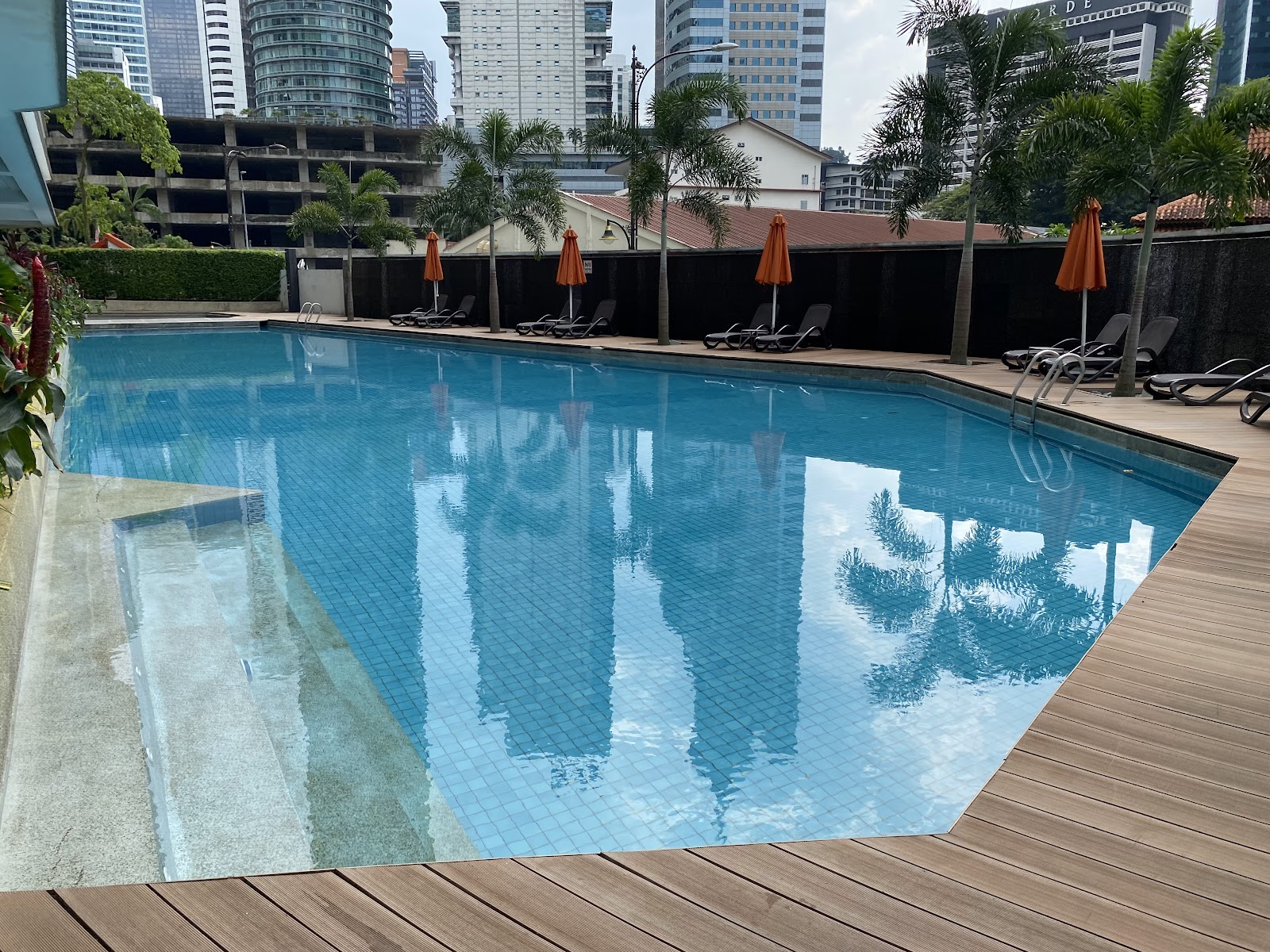 Idaman Residence: A rooftop swimming pool with blue mosaic tiles, surrounded by wooden decking, sun loungers, and closed orange umbrellas, with city buildings in the background.