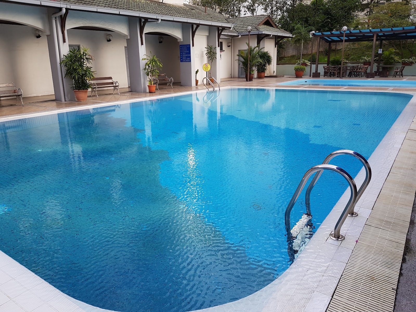 Vista Komanwel B: An outdoor swimming pool with stainless steel ladders, surrounded by a paved deck, benches, and potted plants with a building structure in the background.