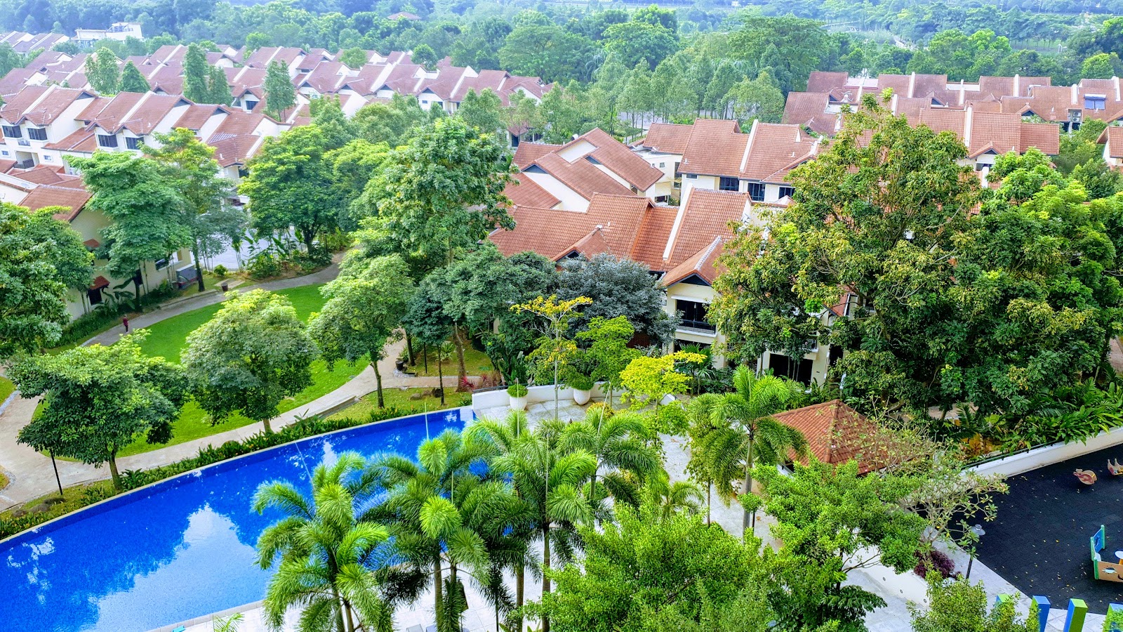 Nadia Parkfront: An elevated view of a residential development with red-roofed houses surrounded by trees, a central green lawn, and a blue swimming pool in the foreground.