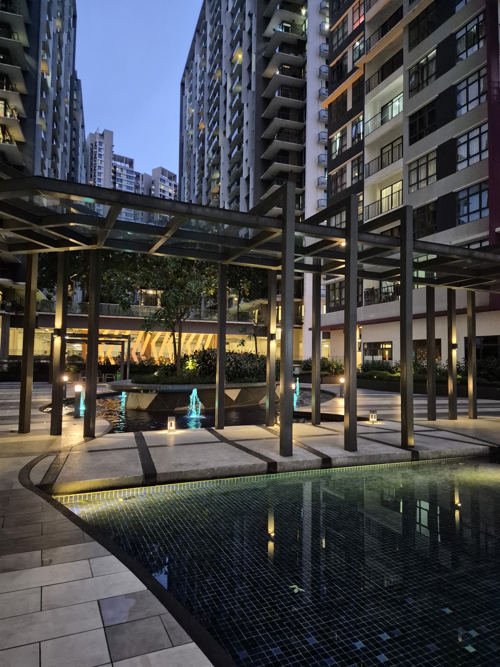 Casa Green: A night view of a residential development's courtyard, featuring a paved walkway, a decorative fountain with blue water jets, a glass-structure pergola, and a reflecting pool in the foreground.