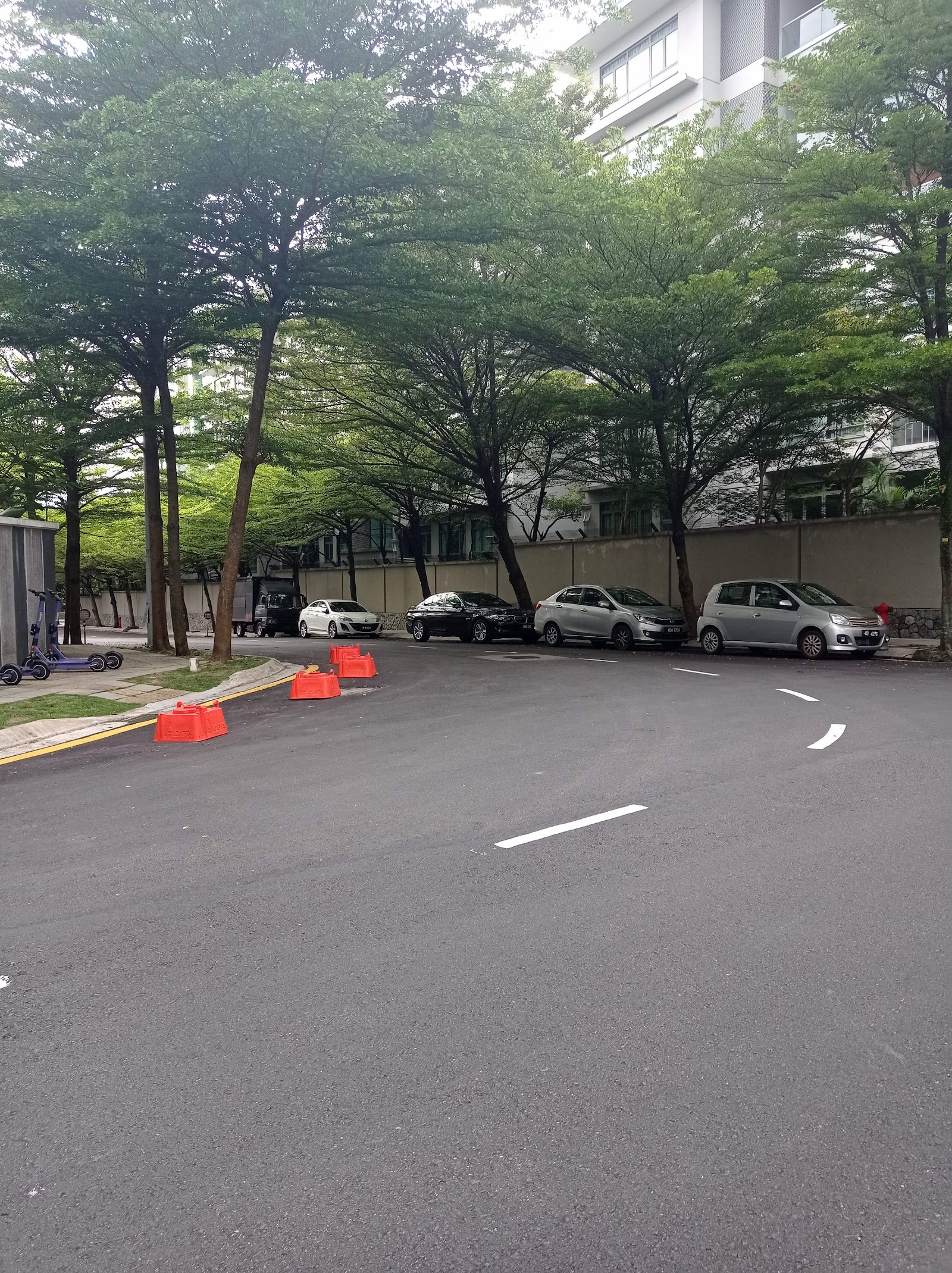 Madge Mansions Condominiums: A paved driveway and car park with several parked vehicles, including a silver hatchback and a black sedan, set among trees with a multi-story building visible in the background.