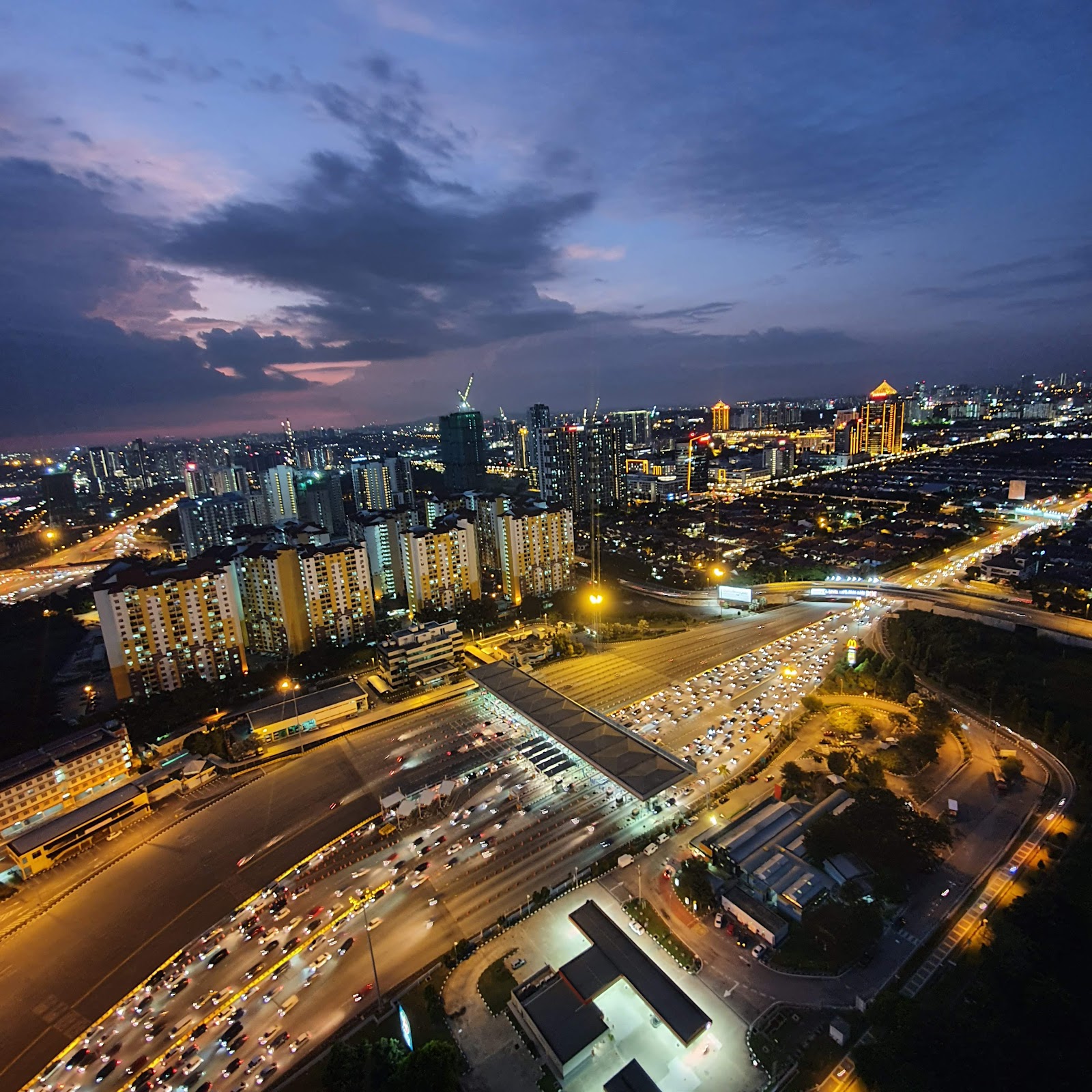 D'Latour: A high-angle aerial view of a city skyline and a highway interchange with heavy traffic during the blue hour.