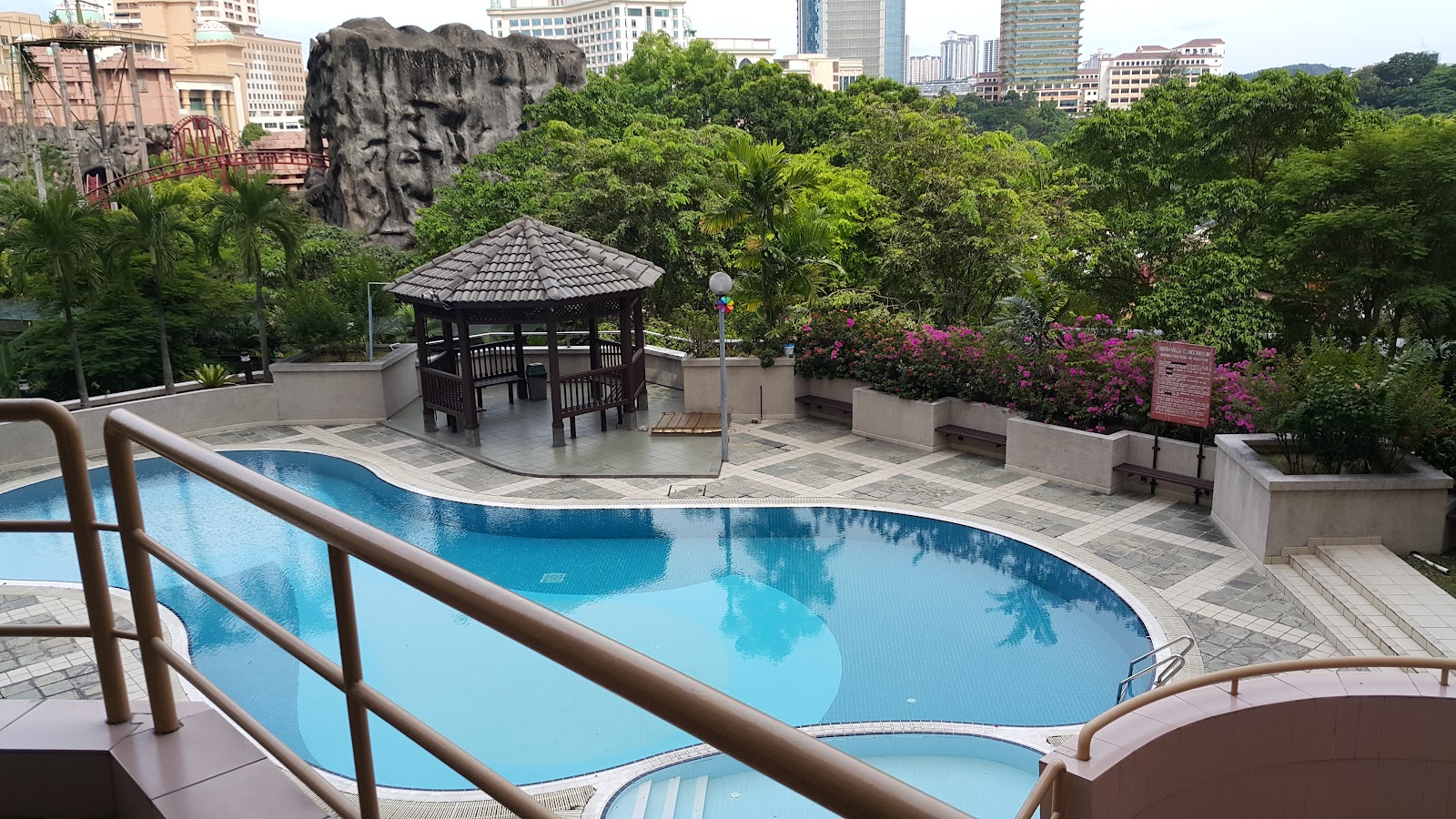 Indah Villa: A swimming pool with a tiled patio, a wooden gazebo, and lush tropical landscaping, including a large artificial rock formation and flowering bushes.