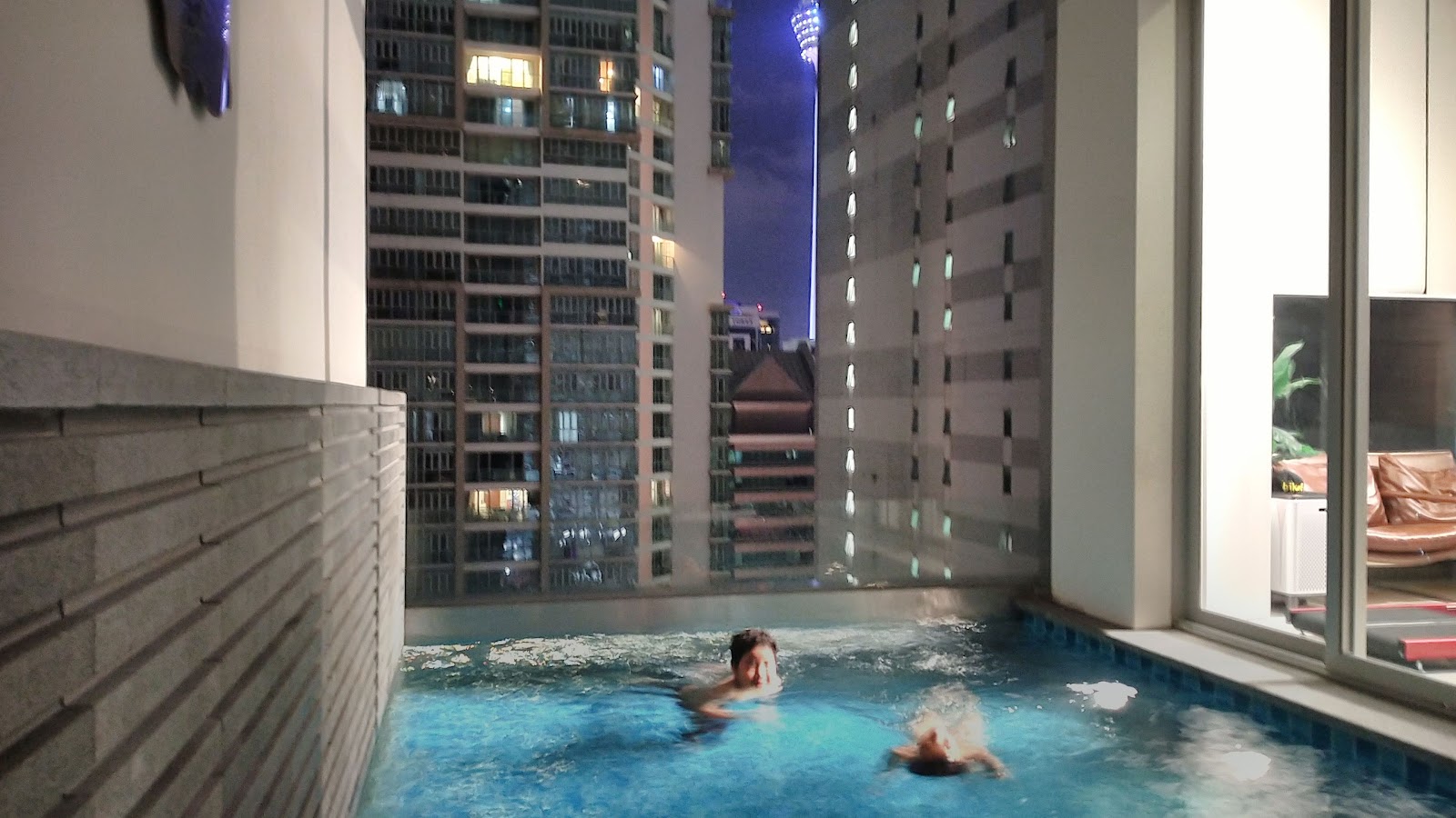 One KL: An outdoor swimming pool on a rooftop or terrace level with people swimming, overlooking a city street and tall buildings at dusk.