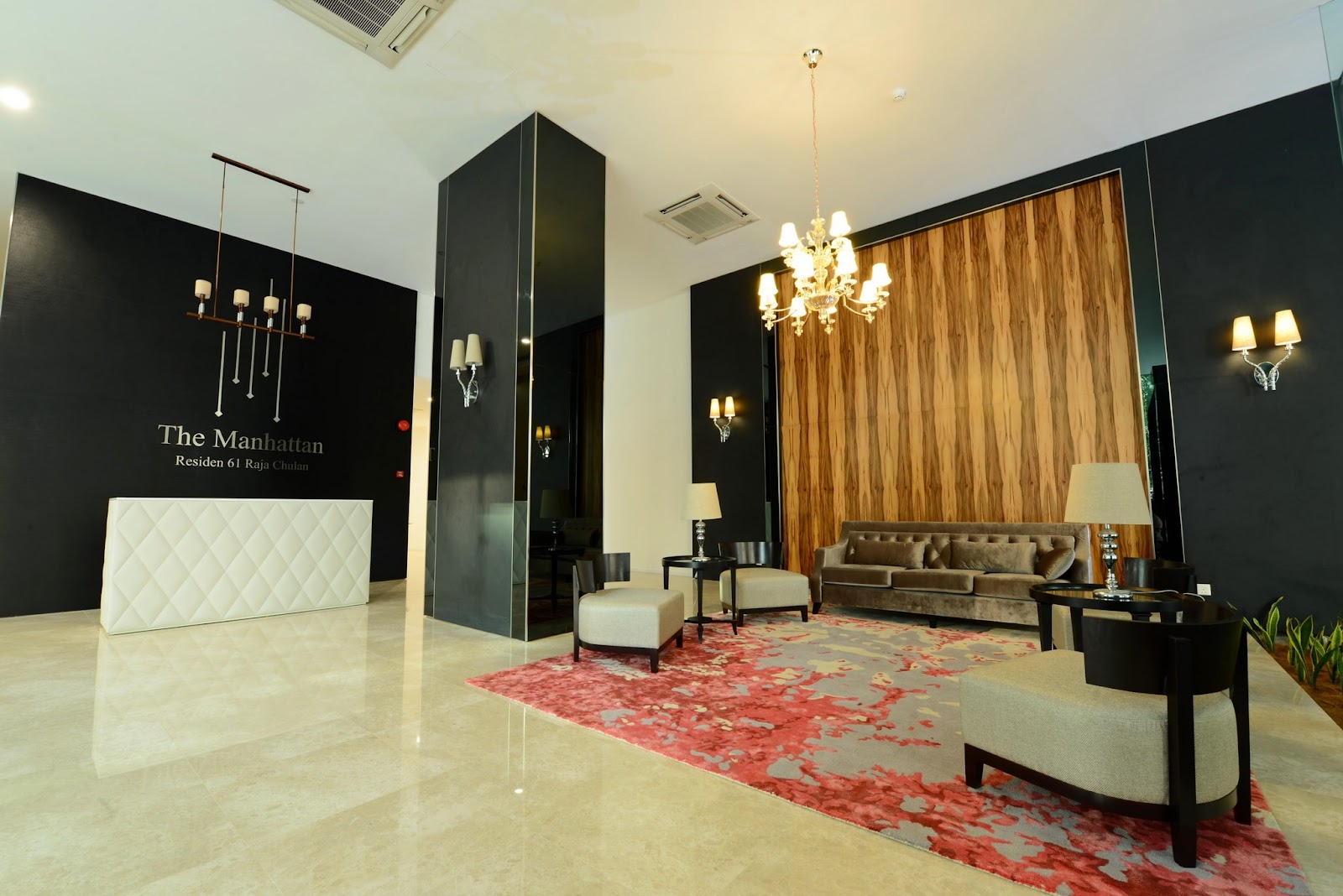 The Manhattan: A modern hotel or apartment lobby featuring a white reception desk, a seating area with a sofa and chairs on a red rug, and a wood-paneled wall.