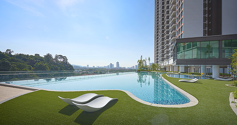 SUNWAY D'HILL RESIDENCES, PETALING JAYA: A long infinity swimming pool on a rooftop with white sun loungers on a green deck, looking out over a landscape with a high-rise building to the right.