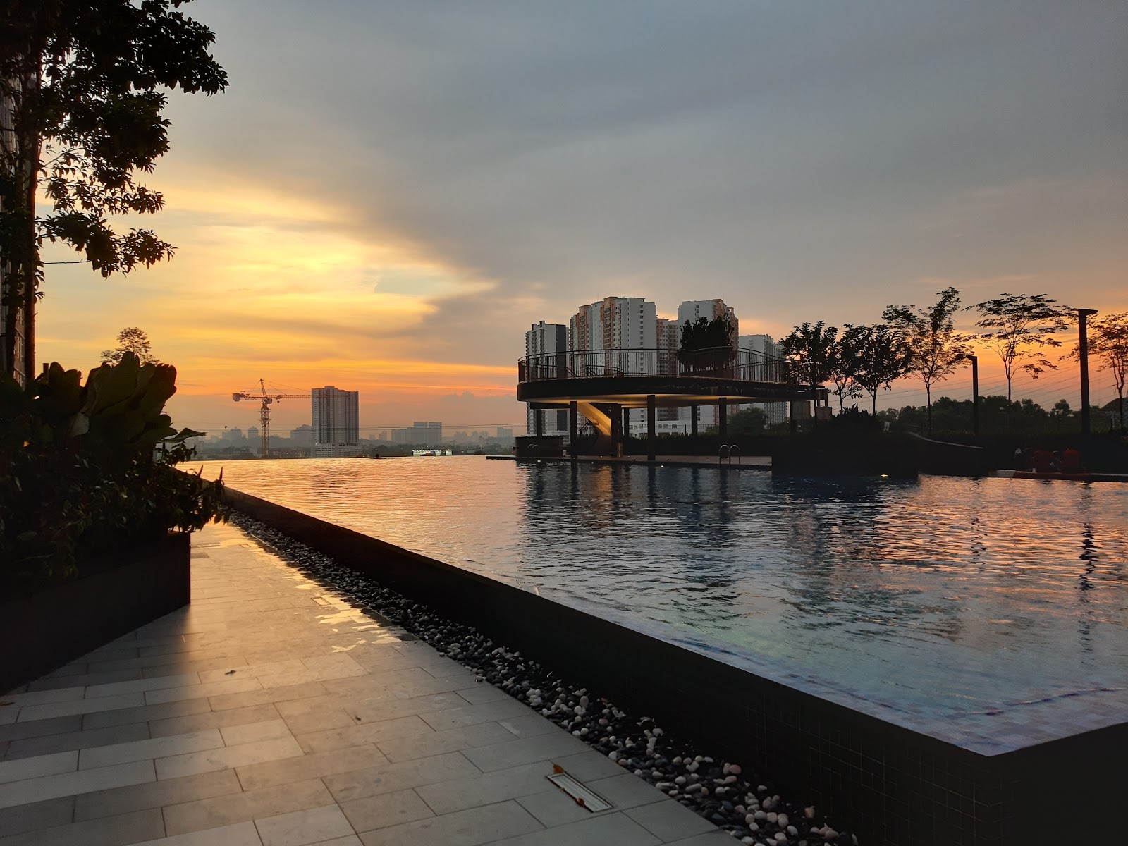 Casa Green: An infinity pool at a residential development at sunset, featuring a circular observation deck in the water and high-rise buildings in the distance.