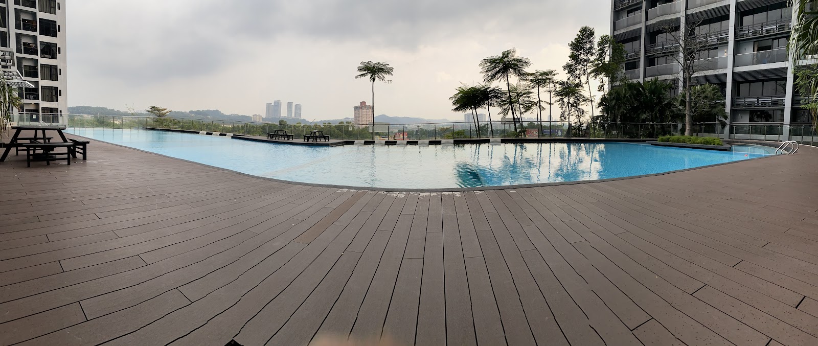 D'Latour: A wide-angle view of an empty, kidney-shaped swimming pool with a blue liner, surrounded by a wooden deck, situated on the rooftop of a residential building with tall buildings and trees in the background.