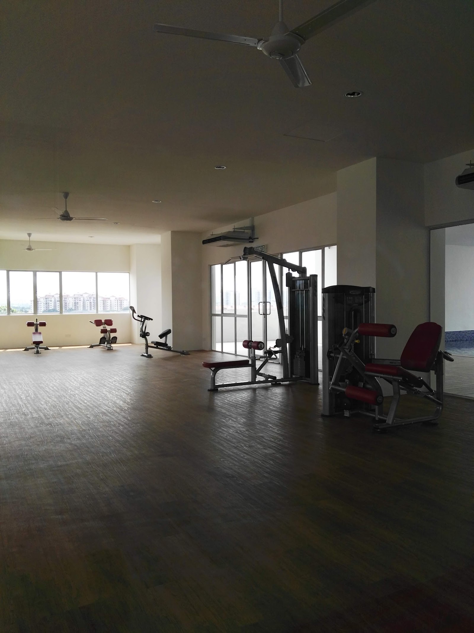 Residensi Puchongmas: An empty indoor gym with weight machines, a squat rack, and an exercise bike on a wood floor, with ceiling fans and large windows.