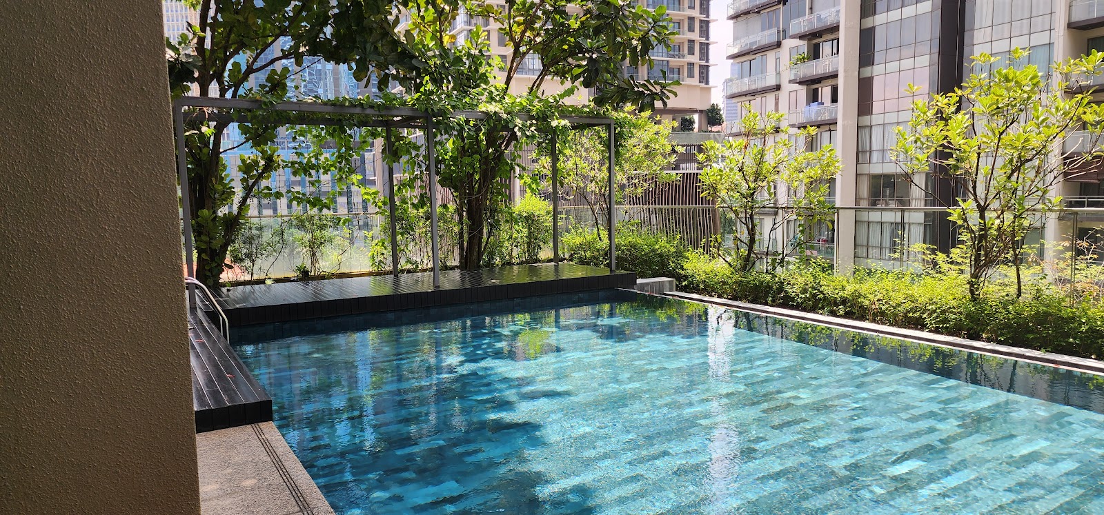 Stonor 3: An outdoor swimming pool with a dark tiled edge and clear water, bordered by green shrubs and trees, with residential apartment buildings visible in the background.