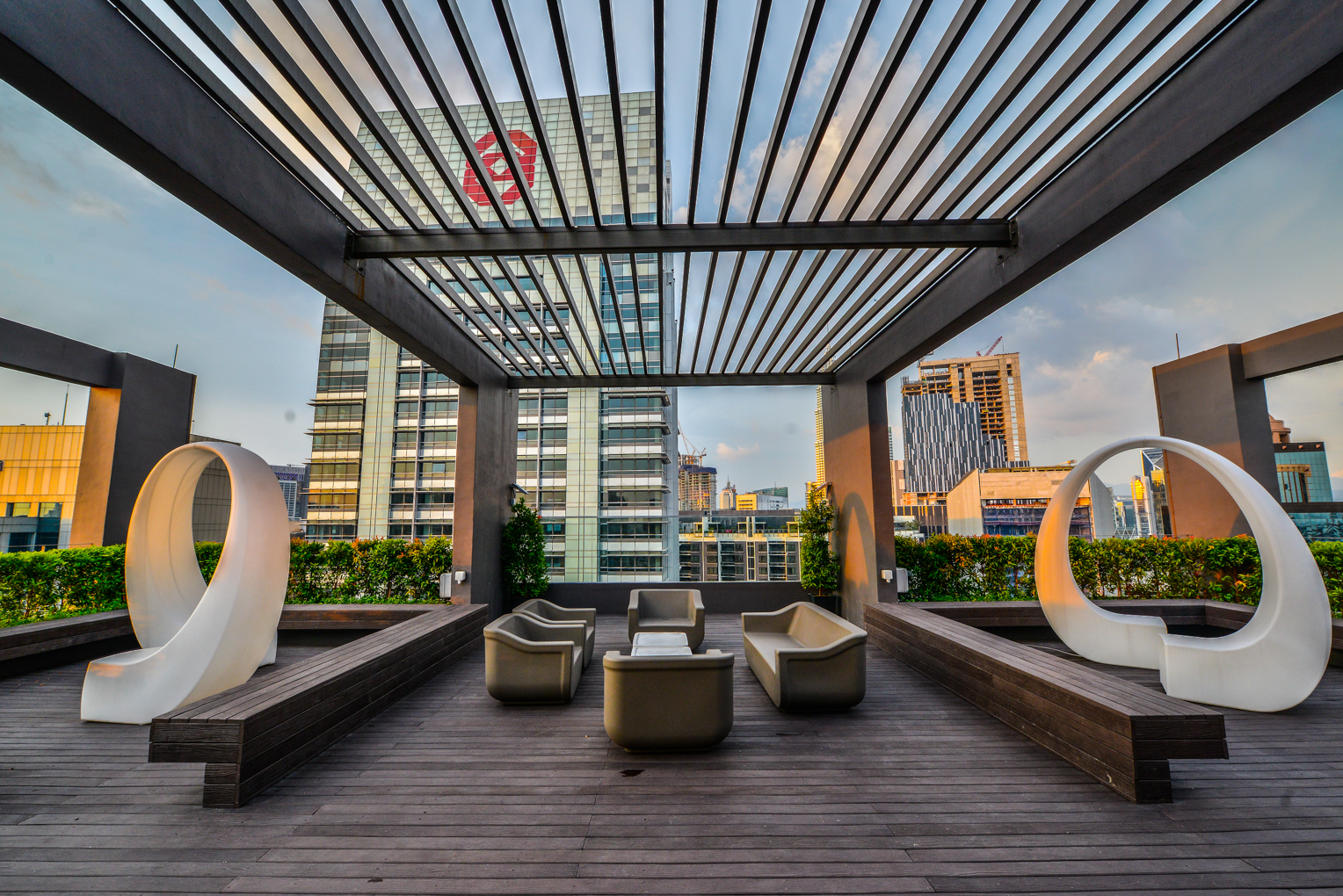 The Manhattan: A furnished outdoor lounge area on a rooftop deck, featuring wooden flooring, seating, and modern white sculptures under a pergola.