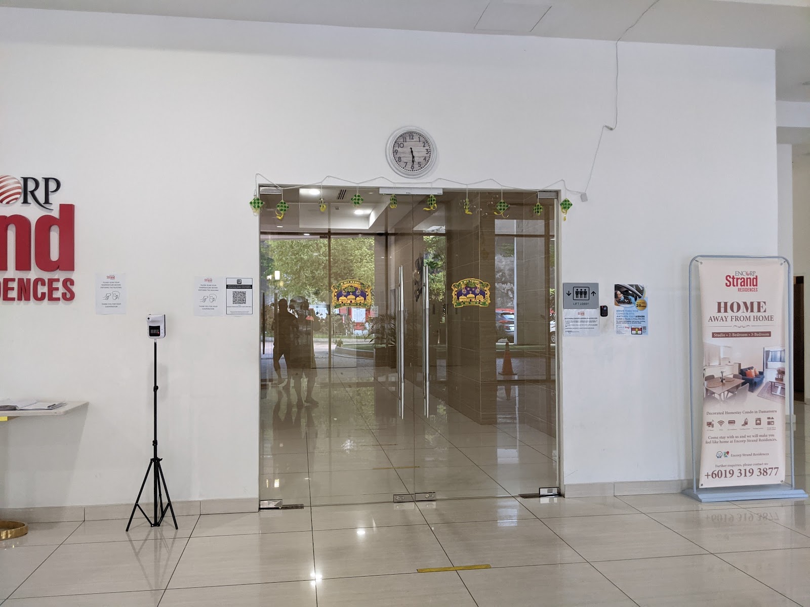Encorp Strand Residences: A view of the building lobby or main entrance, featuring glass doors, a hand sanitizer stand, a wall clock, and a promotional banner.