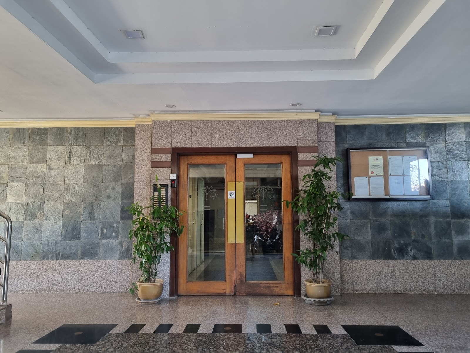 Jelita Court: A lobby entrance with wooden glass double doors, flanked by two potted plants and stone walls, with a notice board on the right.