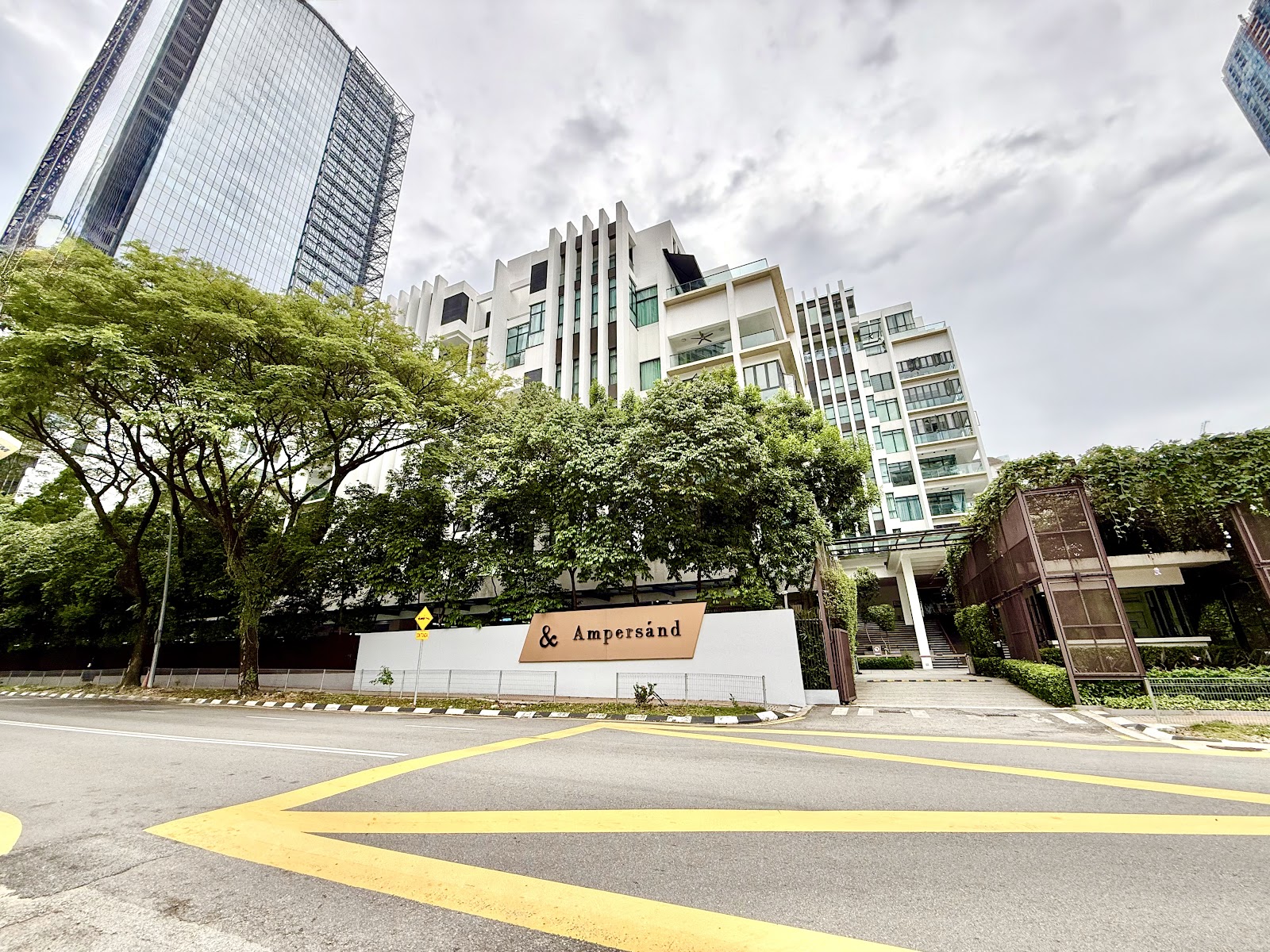 Ampersand: Street-level view of the Ampersand development entrance, showing the white signage wall, the main tower facade, surrounding trees, and a glass-clad skyscraper in the background under an overcast sky.