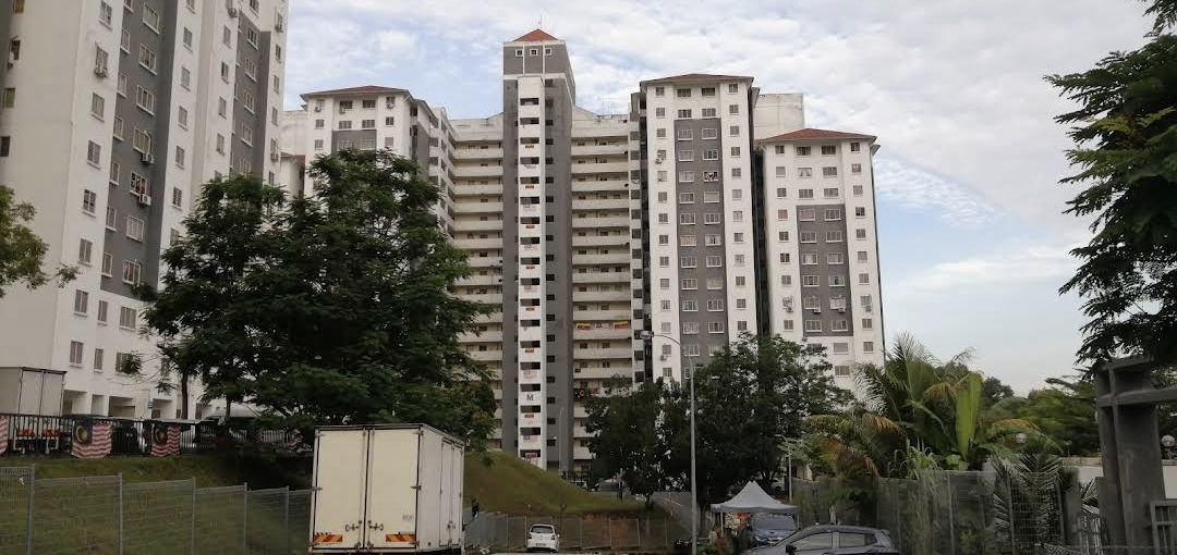 Apartment Seri Ceria 1: A wide shot of a high-rise residential apartment building with white and grey facades, viewed from the ground level near a fenced area and a parked truck.