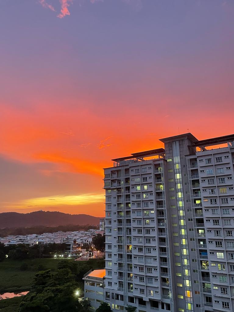 Cova Suites: A resident's view from a high vantage point overlooking a neighboring residential tower, distant hills, and a vibrant sunset sky.