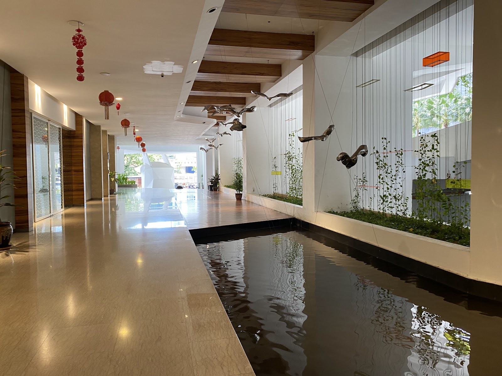 Idaman Residence: A modern interior lobby corridor featuring a reflective water channel, suspended metal bird sculptures, and red lanterns.