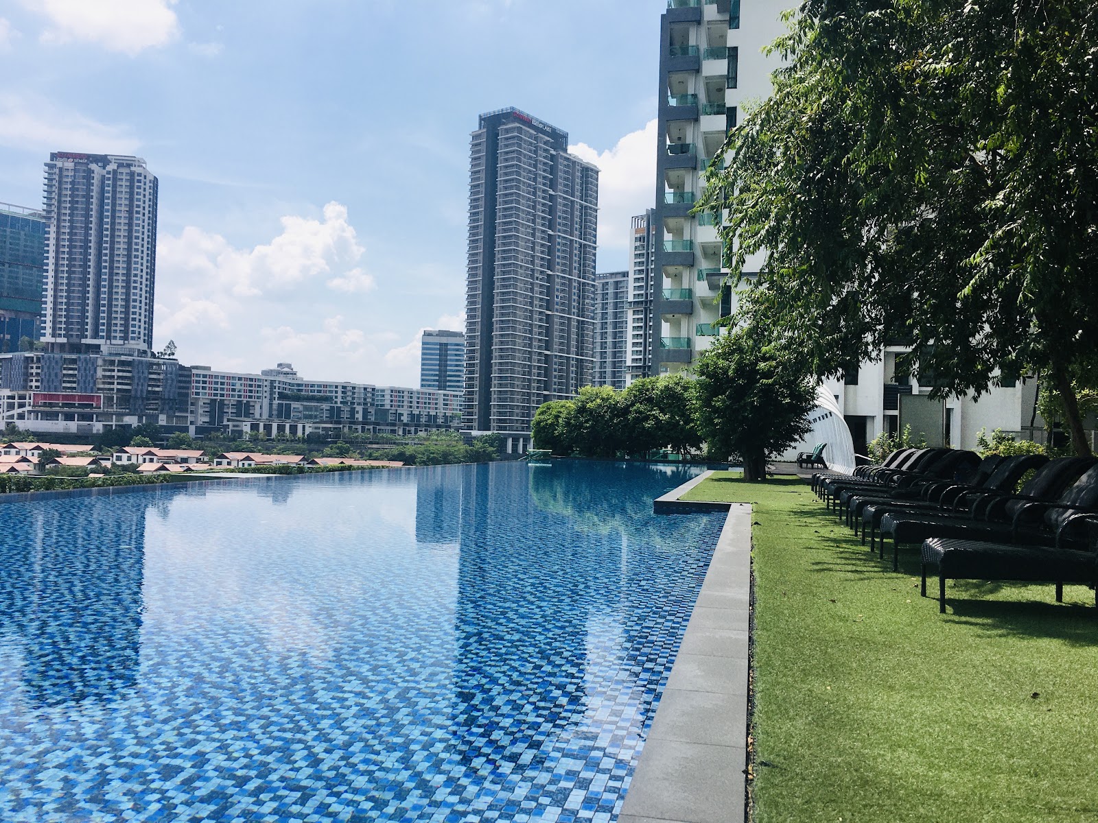 LaCosta, SUNWAY SOUTH QUAY: A view of an outdoor infinity swimming pool with lounge chairs on the grass, looking out towards a cluster of high-rise residential towers.