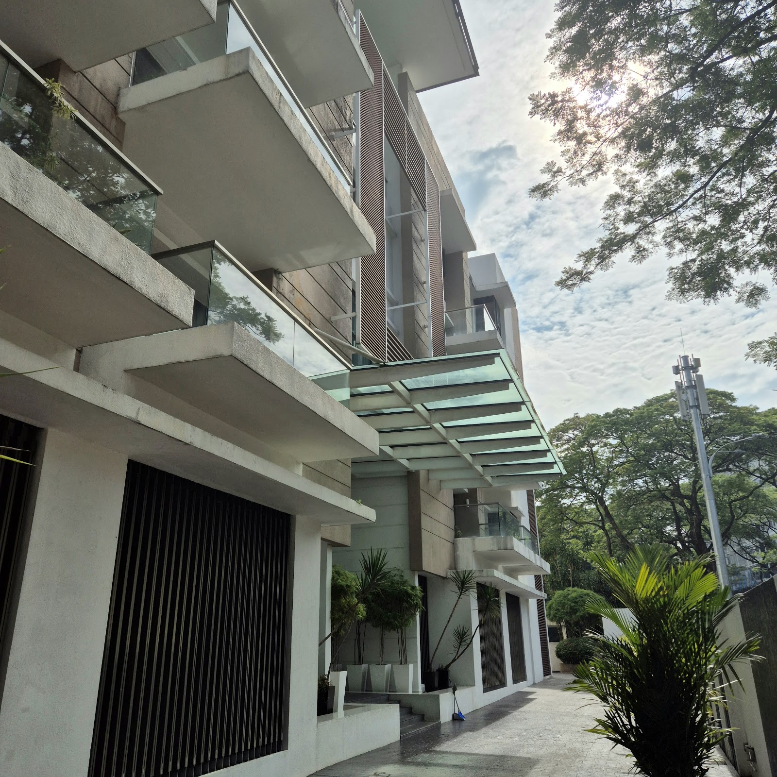 Identiti U-Thant: A low-angle view of the modern exterior facade of a building, featuring concrete balconies, glass railings, and a glass canopy over the ground floor walkway, surrounded by green trees.