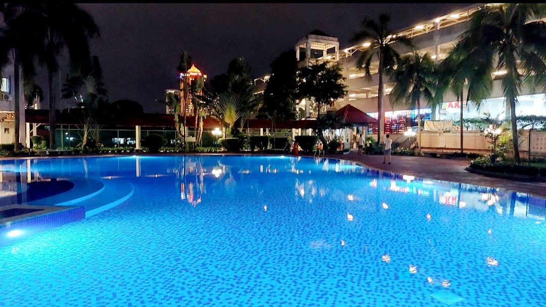 Ridzuan Condominium: A swimming pool illuminated by underwater blue lights at night, set within the landscaped grounds of the condominium with palm trees and a building in the background.